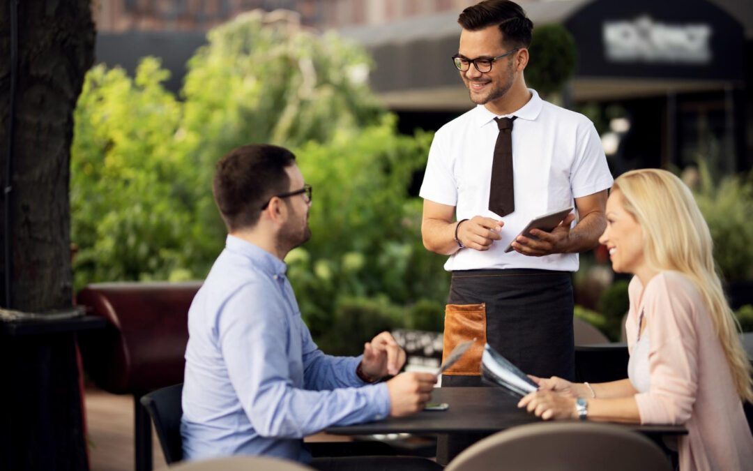 Serveur souriant prenant une commande avec un pad numérique auprès de deux clients en terrasse dans un restaurant moderne.